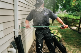 A uniformed and masked dryer vent cleaning pro cleans a duct line from the exterior of house.